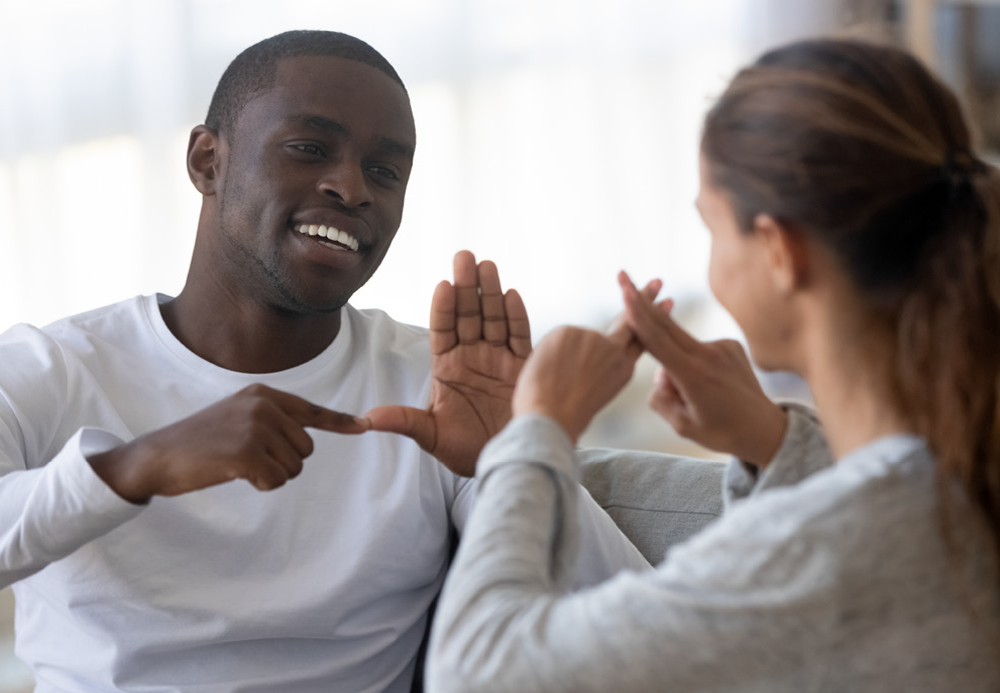 lack man in white crew neck, smiling, and signing to woman in white crew neck