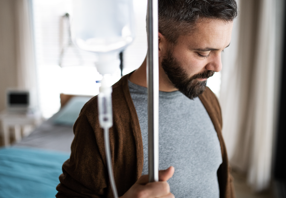 Man with dark and hair and a beard, looking down, standing with an IV drip