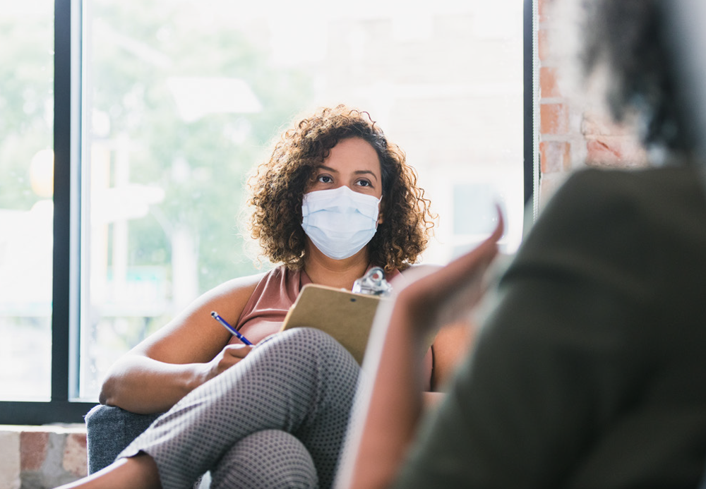 A woman with a mask and casually dressed, sitting and holding a chart, talking to an older woman sitting across from her