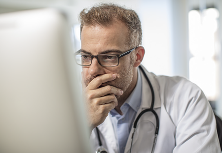 A man in a wight lab coat contemplating while viewing a computer monitor