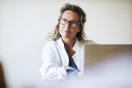 A seated scientist listens intently to a person out of frame as she takes notes in her laptop