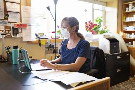 A woman wearing a mask sits at her desk in her office while working on her computer