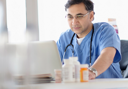 A doctor searches his laptop with 3 bottles of pill in the foreground of the frame