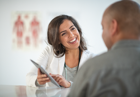 A smiling nurse shows a patient sitting across from her something on the tablet she is holding