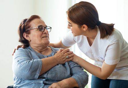 Seated elderly woman touches the hand of her caretaker who has one arm around her