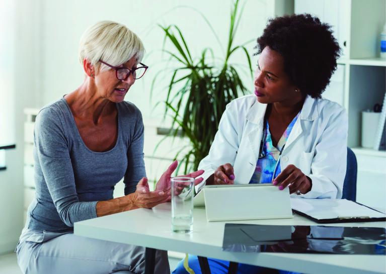 woman with short hair and glasses sitting at a table with a healthcare provider in lab coat sitting next to her having a conversation